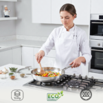 Female chef in white chef's coat sautéing colourful mixed vegetables in a stainless steel Pro Fry Pan on a gas hob in a modern white kitchen, with TriPly, Eco Friendly, and induction compatibility icons displayed at the bottom